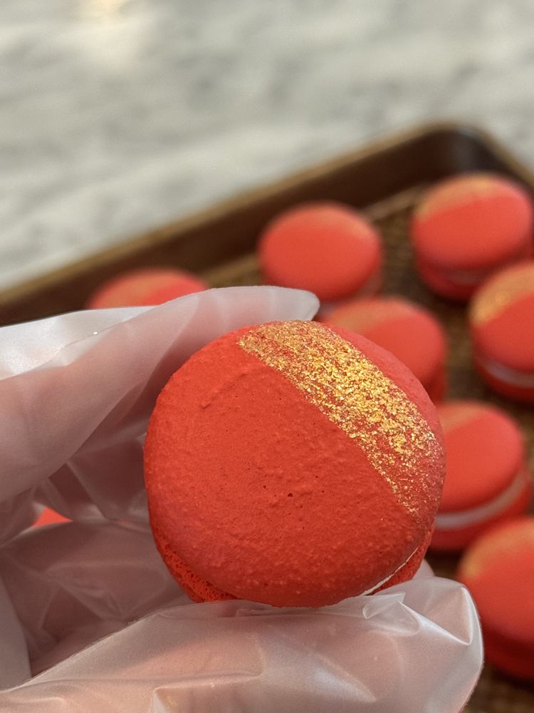 A hand holding a red macaron shell brushed with gold, with more red shells on a tray behind