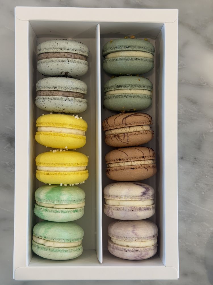 A white window gift box holding twelve macarons in green, yellow, brown, and lavender